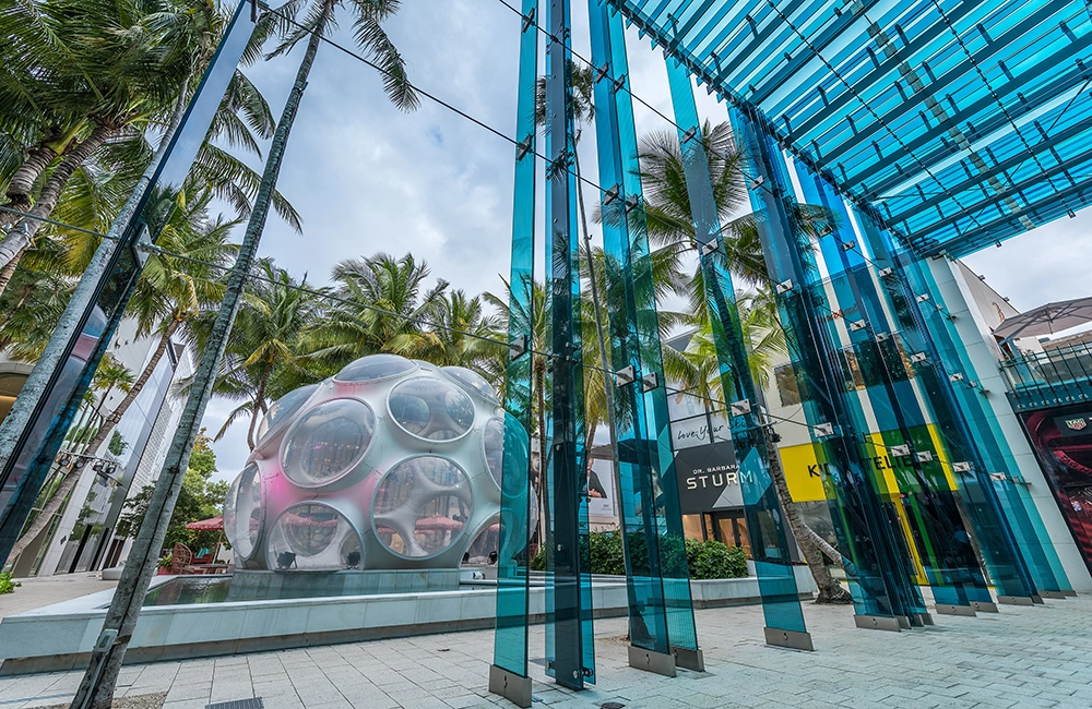 Blue glass architectural panels frame a modern plaza with palms and a sculptural pavilion in Miami