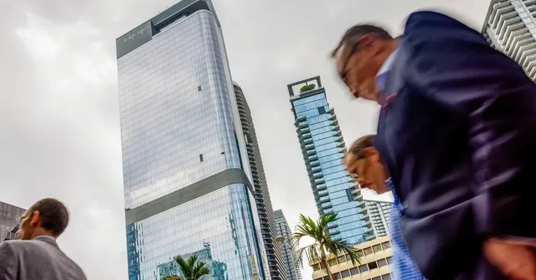 Businesspeople walk past gleaming glass skyscrapers and luxury high-rise towers in a thriving urban downtown real estate district.