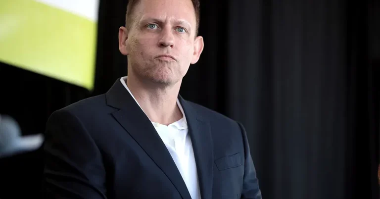 A man in a navy blazer and white polo shirt sits attentively during a professional presentation event indoors.